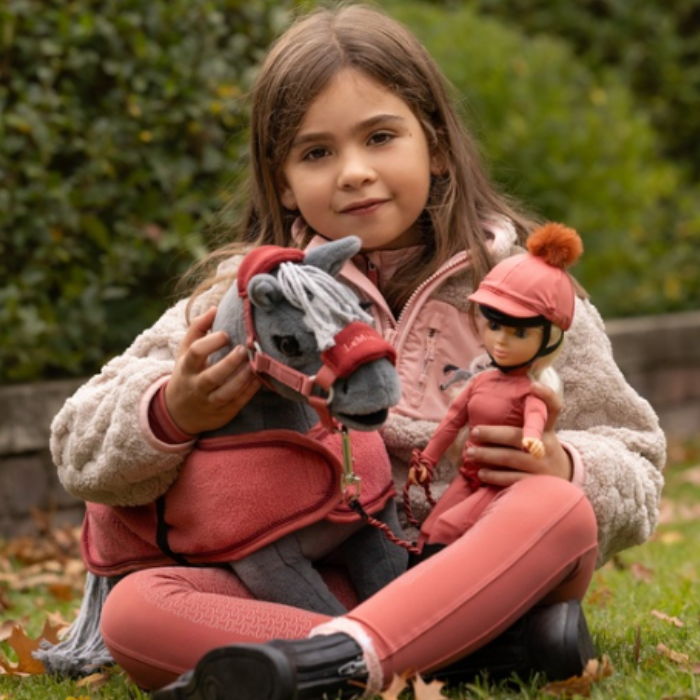 LeMieux AW26 Toy Rider Outfit Varient Spiced. Young girl holding a mini figure and toy rider wearing a matching spiced outfit sat in a garden. 