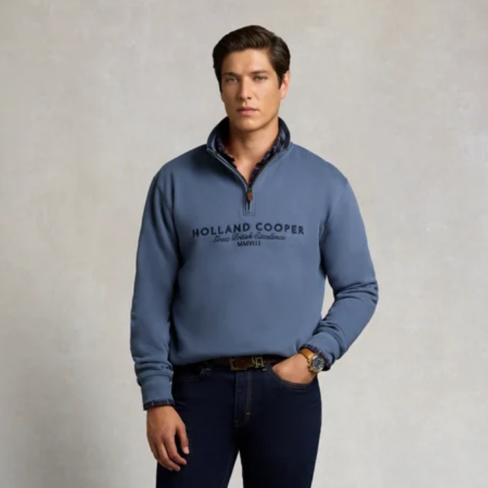 Holland Cooper Heritage Quarter Zip Sweat (Men's) Variant Denim Blue. Man wearing a blue 1/4 zip sweatshirt with Holland Cooper branding across the chest in navy agaisnt a grey background. 