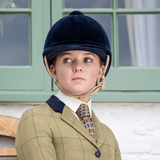 Person wearing a navy riding helmet and tweed jacket with a patterned tie, standing in front of a green window.