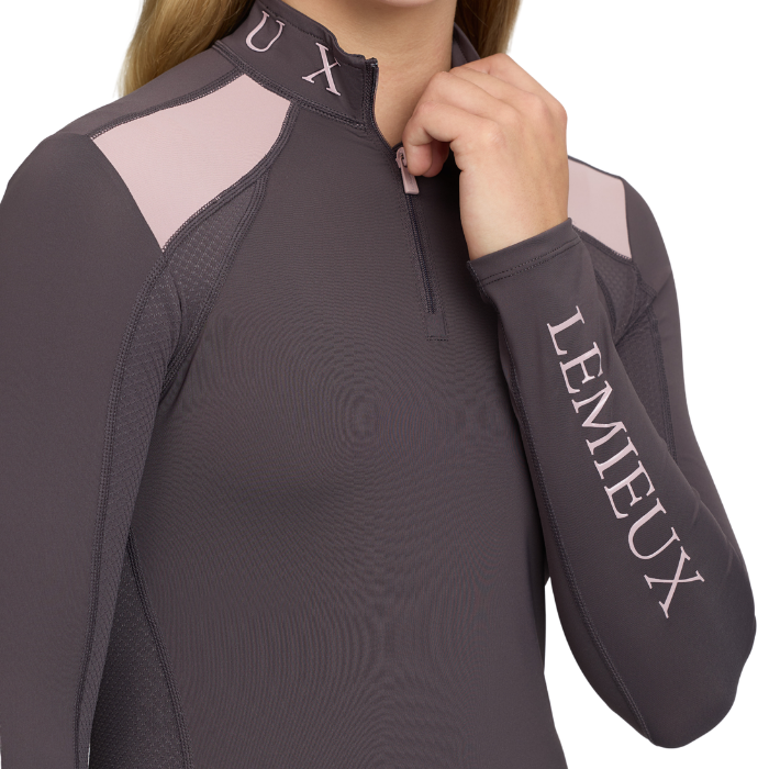 LeMieux AW26 Young Rider Mia Mesh Base Layer Varient Cocoa. Close-up of a woman wearing a dark gray long-sleeve quarter-zip athletic top with light pink shoulder panels and 'LEMIEUX' printed in white down the right sleeve.