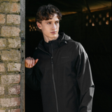 LeMieux AW26 Mens Waterproof Lightweight Jacket Varient Black. A man wearing a black hooded zip-up jacket stands in a doorway next to a brick wall, looking off-camera. The jacket has a subtle tonal logo on the left chest.