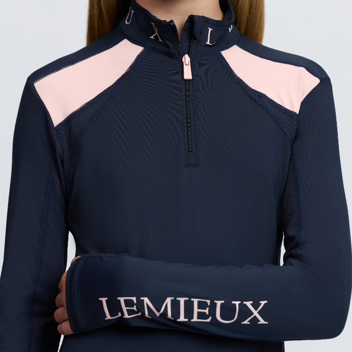 LeMieux AW26 Young Rider Mia Mesh Base Layer Varient Navy. A close-up of a model wearing a navy blue long-sleeve athletic quarter-zip top with light pink shoulder panels and 'LEMIEUX' embroidered in pink on the left sleeve.