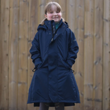 A young girl wearing a dark navy hooded parka coat with a green shirt underneath, standing in front of a wooden fence.