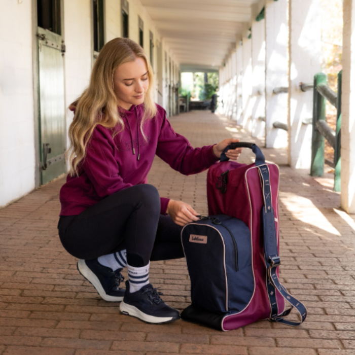 LeMieux AW26 Boot & Hat Bag Varient Blackberry. A woman crouched in an equestrian stable corridor, organizing a navy and blackberry equestrian hat and boot bag whilst holding the top handle. 