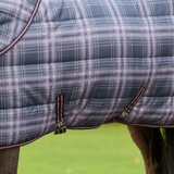 Close-up of a quilted horse blanket with a blue-gray and lavender plaid pattern, featuring dark brown trim and metal snap fasteners, shown on a horse with green grass in the background.