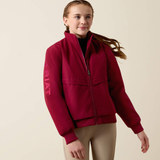 A young girl wearing a burgundy zip-up jacket with 'ARIAT' printed in bright red on the sleeve, paired with beige riding pants, against a solid light beige background.