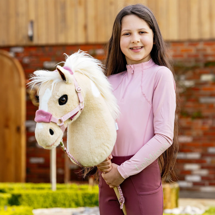 LeMieux AW26 Hobby Horse Vogue Headcollar & Leadrope Variant Colour Blossom. 
A young person with a plush toy horse head mounted on a stick, with a white body, white faux-fur mane, and blossom colour halter, photographed outdoors against a blurred garden background.