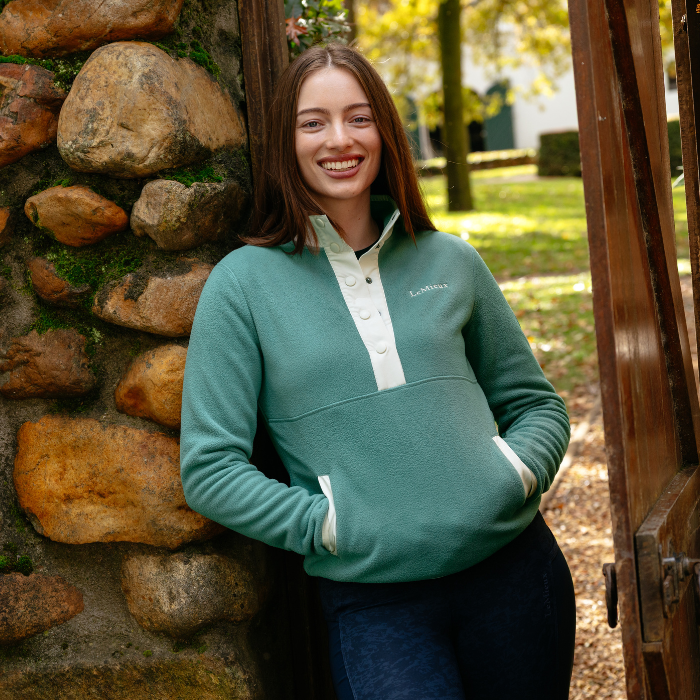 LeMieux AW26 Piper Popper Fleece Varient Aloe. A woman smiling and leaning against a rustic stone wall and wooden gate, wearing a sage green quarter-zip pullover with a white placket and white pocket trim. The brand name 'LeMieux' is visible on the chest.
