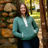 LeMieux AW26 Piper Popper Fleece Varient Aloe. A woman smiling and leaning against a rustic stone wall and wooden gate, wearing a sage green quarter-zip pullover with a white placket and white pocket trim. The brand name 'LeMieux' is visible on the chest.