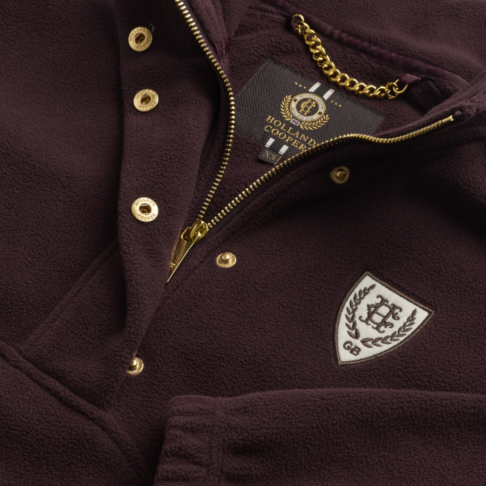 Holland Cooper Cranborne Over Head Fleece Variant Mulberry. Close-up of a dark plum fleece pullover jacket with gold-tone snap buttons, a gold zipper, a brand label reading Holland Company, and a white shield patch on the chest.