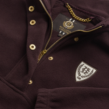 Holland Cooper Cranborne Over Head Fleece Variant Mulberry. Close-up of a dark plum fleece pullover jacket with gold-tone snap buttons, a gold zipper, a brand label reading Holland Company, and a white shield patch on the chest.