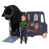 LeMieux AW26 Toy Pony Lorry Wooden Varient Brown. A doll with long red hair stands in front of a horse trailer and black horse. The doll wears a green long-sleeve top, blue pants, and black boots, and holds a rope leading to the horse.