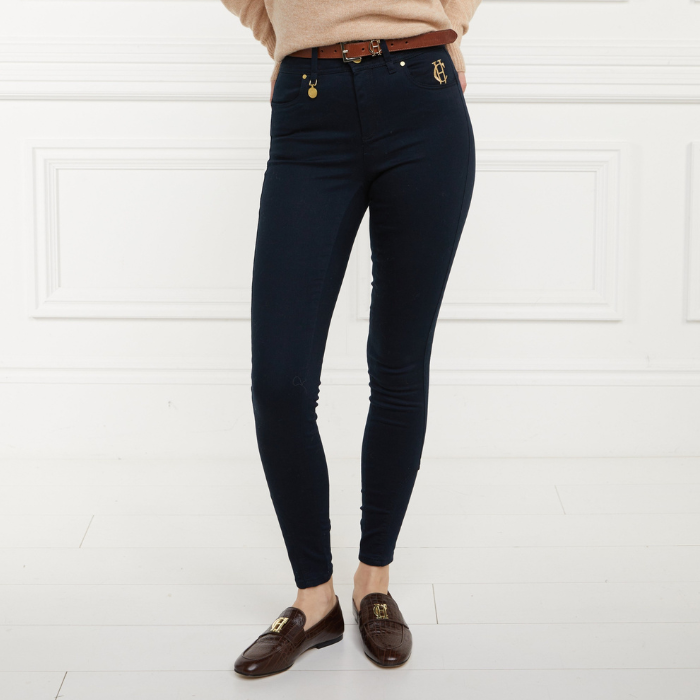 Holland Cooper Jodhpur Jean Variant Ink Navy. A person wearing slim-fit dark navy skinny jeans with gold-tone hardware, paired with a tan sweater and brown loafers, standing against a white paneled wall.