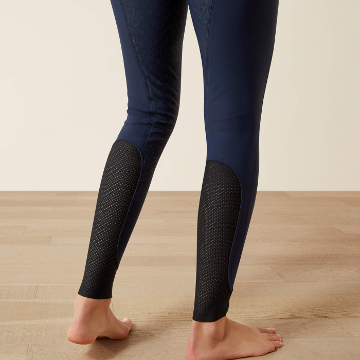 A person wearing navy blue leggings with black textured mesh knee and calf panels, standing barefoot on a light wood floor against a beige wall.
