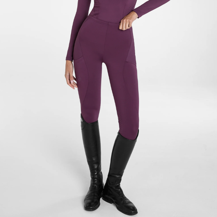 Aztec Diamond AW26 Core Leggings Full Seat Variant Plum. A model wearing high-waisted, form-fitting equestrian breeches in a deep plum purple, paired with black riding boots, shown from the waist down on a light gray studio background.