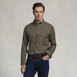Holland Cooper Men's Classic Fit Brushed Cotton Shirt Variant Ruby Khaki Check. A man wearing a brown and beige plaid long-sleeve button-down shirt with dark blue jeans, standing against a textured light gray background.