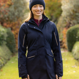 LeMieux AW26 Stormwear Performance Riding Coat Varient Navy. A woman wearing a navy blue hooded jacket with contrast zippers, a dark knit beanie, and a burgundy top, standing in a garden setting.