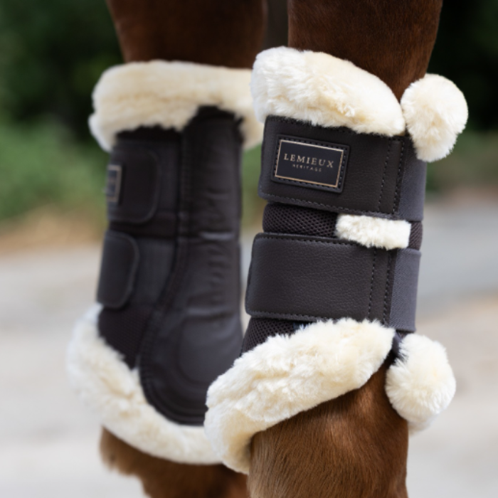 LeMieux AW26 Heritage Fleece Edged Brushing Boot Varient Cocoa. Close-up of a horse's lower legs wearing brown protective boots with thick cream-colored fleece lining. A brand label reading 'LeMieux Heritage' is visible on the front boot.