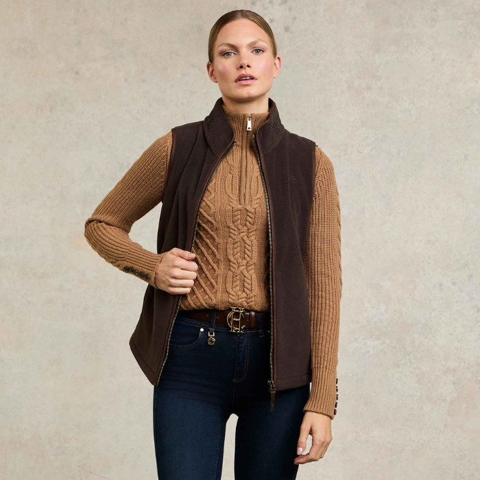 Holland Cooper Country Fleece Gilet Variant Chocolate. Woman wearing a chocalte gilet with brown detailing, over an orange jumper with blue jeans against a grey background. 