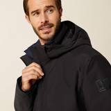 A man wearing a black Ariat hooded jacket, shown from the chest up against a plain light beige studio background. The Ariat logo patch is visible on the left sleeve.