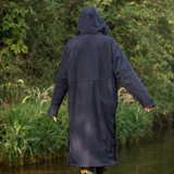 A person wearing a long navy hooded raincoat stands outdoors by a body of water with green reeds and foliage in the background, photographed from the back.