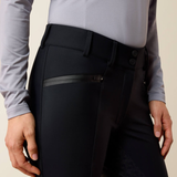 Close-up of a person wearing black equestrian riding breeches with a zippered pocket and a light gray long-sleeve shirt, against a plain beige background.