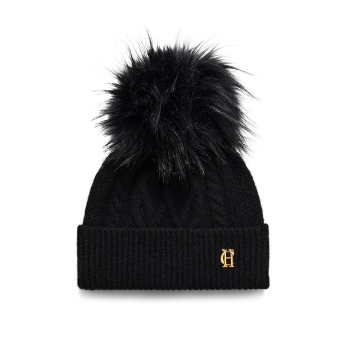 Holland Cooper Sloane Cable Bobble Hat Variant Black. A black cable-knit beanie with a large black faux fur pom-pom and a small gold embroidered monogram logo on the front cuff, shown against a plain white background.