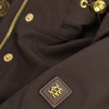 Holland Cooper Essential Waterproof Jacket Variant Bournville. Close-up of a dark brown jacket or coat fabric with gold-tone hardware, including a zipper, buttons, and a small square patch with a gold crest logo.
