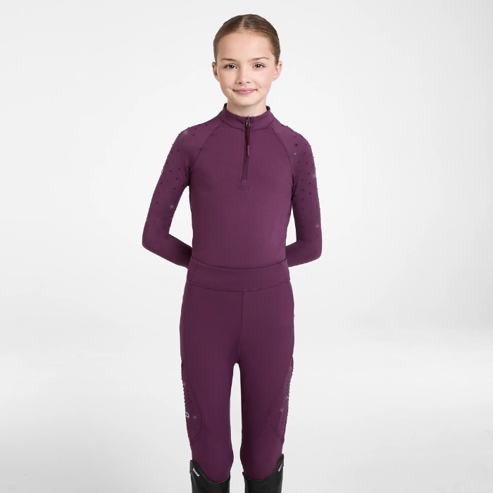 A young girl stands against a light gray studio background wearing a matching long-sleeve half-zip athletic top and leggings in a deep plum purple color, paired with black boots.