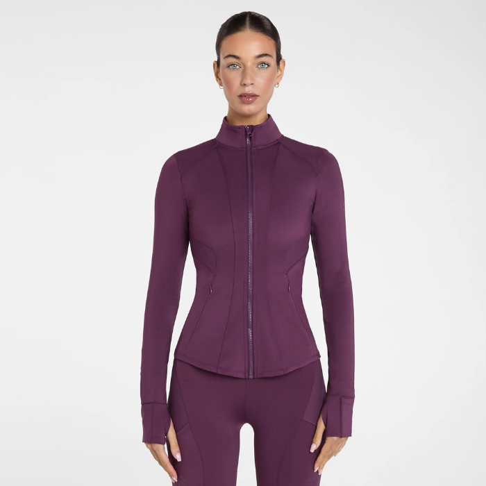Aztec Diamond AW26 Core Fitted Jacket Variant Plum. A woman modeling a fitted long-sleeve athletic zip-front jacket in a deep plum purple color, paired with matching leggings, against a clean light grey studio background.
