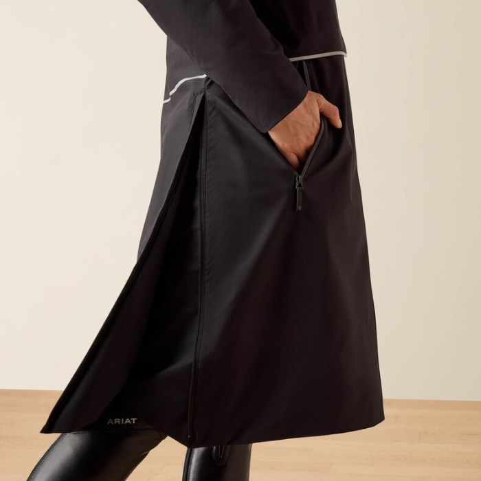 A close-up of a person wearing a black Ariat coat with a side zipper, shown from the waist down against a neutral beige background.