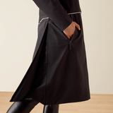 A close-up of a person wearing a black Ariat coat with a side zipper, shown from the waist down against a neutral beige background.