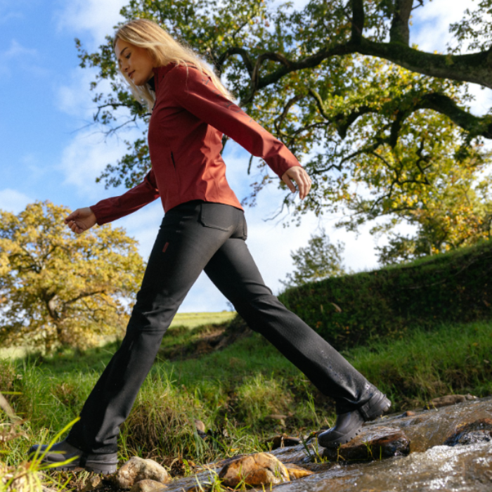 LeMieux AW26 Stormwear Stretch Waterproof Trousers Varient Black.  A woman hiking outdoors on a rocky stream, wearing a rust-red long-sleeve jacket and black pants.