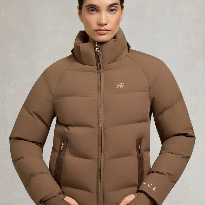 Holland Cooper Chiltern Yard Jacket Variant Chestnut. A woman wearing a brown puffer jacket with a high collar, front zipper, and zippered hand pockets, photographed against a neutral gray studio background.