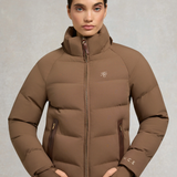 Holland Cooper Chiltern Yard Jacket Variant Chestnut. A woman wearing a brown puffer jacket with a high collar, front zipper, and zippered hand pockets, photographed against a neutral gray studio background.
