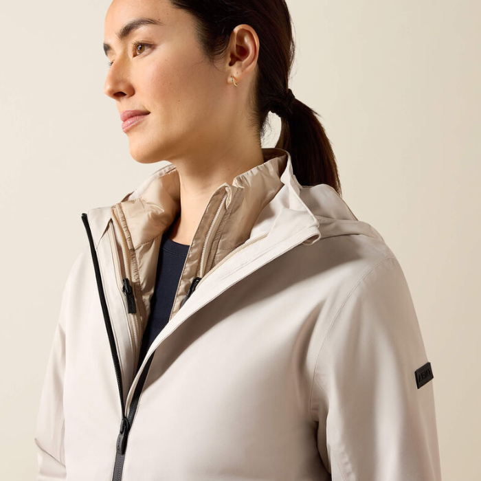 A woman in profile wearing a light beige hooded jacket with a black zipper and a small black sleeve label, photographed against a plain light beige background.
