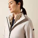 A woman in profile wearing a light beige hooded jacket with a black zipper and a small black sleeve label, photographed against a plain light beige background.