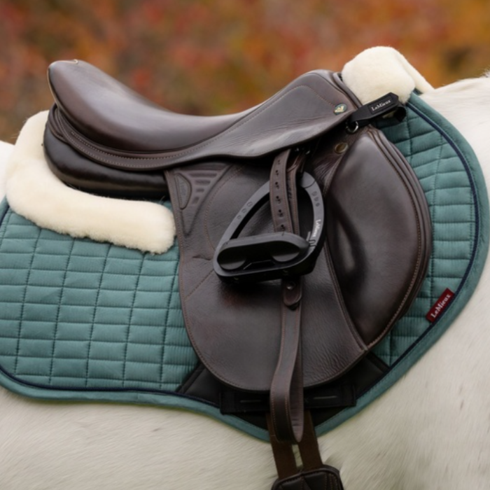 LeMieux AW26 Merino+ Suede Classic Close Contact Square Varient Aloe. A black leather equestrian saddle resting on a Aloe quilted horse pad with white fleece lining, shown on a horse against a blurred outdoor background.