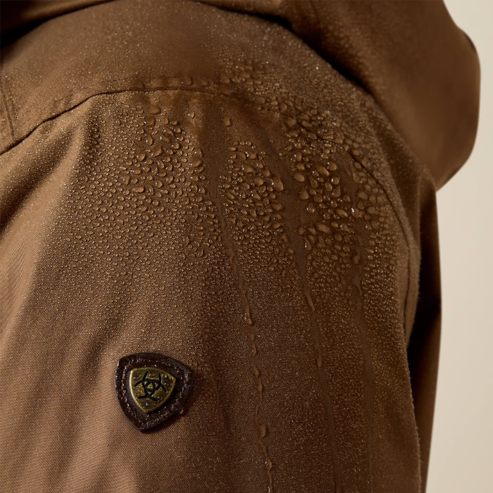 Close-up of a brown waterproof jacket with water droplets beaded on the fabric and a small shield-shaped logo patch on the shoulder.
