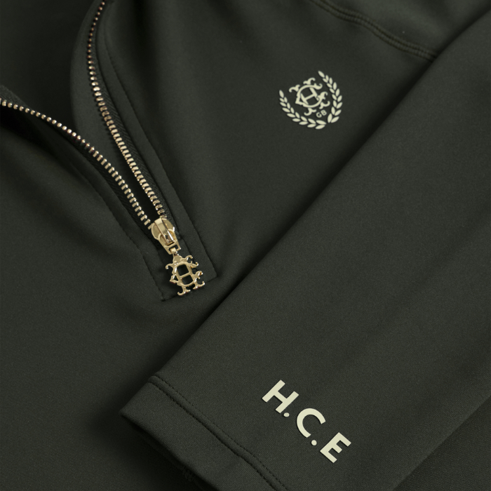 A Holland Cooper Cranborne Long Sleeve Base Layer Variant Colour Dark Olive. 
Close-up of a dark olive green zip-up jacket featuring a gold-tone zipper with a decorative crest pull, a matching gold crest embroidered on the chest, and 'H.C.