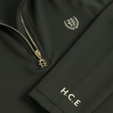 A Holland Cooper Cranborne Long Sleeve Base Layer Variant Colour Dark Olive. 
Close-up of a dark olive green zip-up jacket featuring a gold-tone zipper with a decorative crest pull, a matching gold crest embroidered on the chest, and 'H.C.