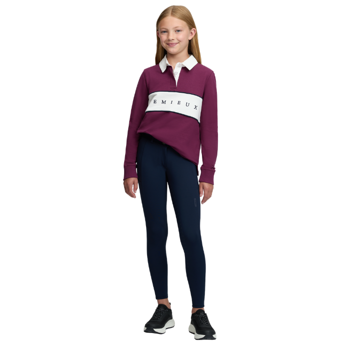 LeMieux AW26 Young Rider Riley Rugby Shirt Varient Blackberry. A young girl modeling a long-sleeve rugby-style top in deep burgundy with a white collar and a white horizontal chest stripe featuring the brand name 'LEMIEUX', paired with navy leggings and black sneakers, against a plain white background.