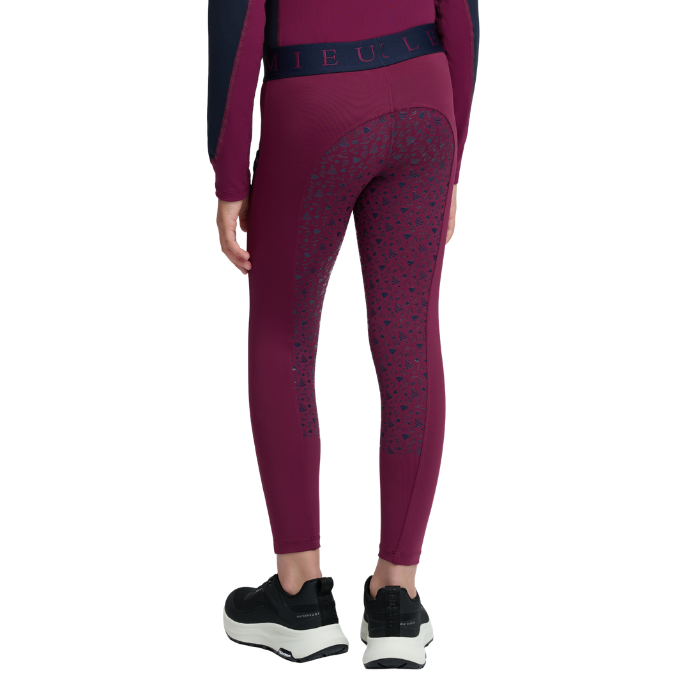 LeMieux AW26 Mini Pull Ons Varient Blackberry. Rear view of a model wearing burgundy athletic leggings with dark navy patterned panels and a matching waistband, paired with black and white sneakers on a white background.