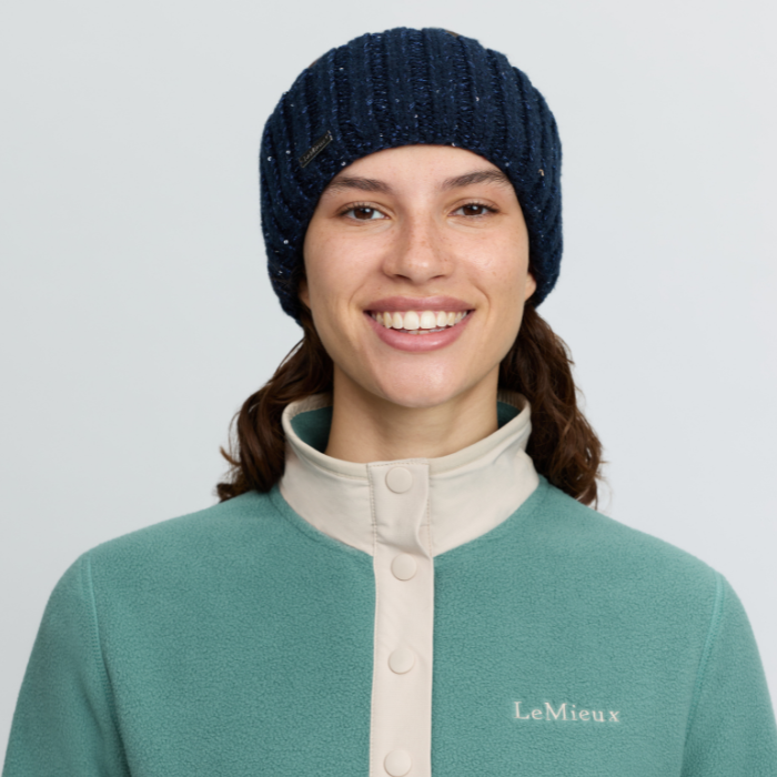 LeMieux AW26 Bernie Sequin Headband Variant Colour Navy.
A smiling woman wearing a dark navy blue knit beanie and a teal green fleece pullover with a cream collar and buttons. The brand name 'LeMieux' is embroidered on the left chest.