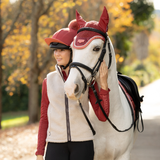 LeMieux AW26 Young Rider Trinity Teddy Gilet Varient Almond. A woman in equestrian apparel stands beside a white horse outdoors on a path with autumn trees in the background. The horse is wearing a red fly veil and black bridle, while the woman wears a cream fleece vest over a red long-sleeve top and black breeches.
