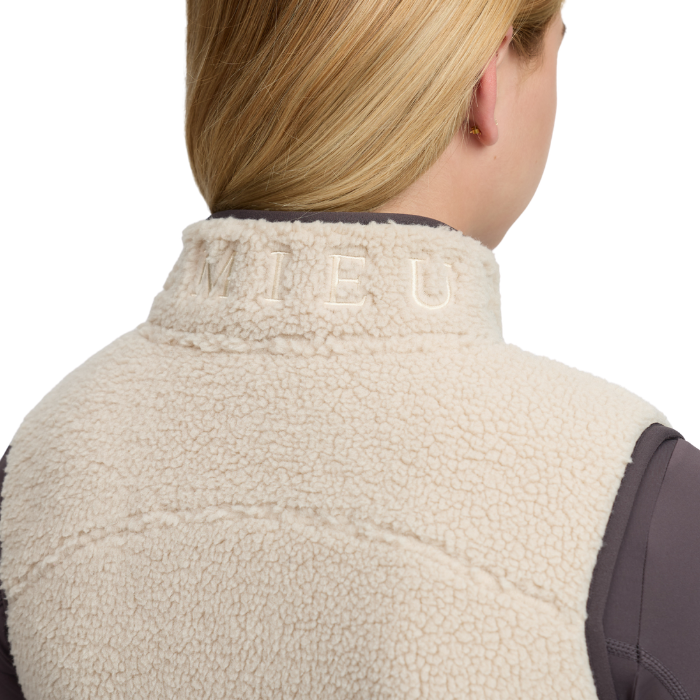 LeMieux AW26 Young Rider Trinity Teddy Gilet Varient Almond. Close-up back view of a person wearing a cream sherpa fleece vest over a dark gray top, with the brand name LEMIEUX embroidered on the collar.