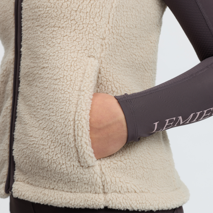 LeMieux AW26 Young Rider Trinity Teddy Gilet Varient Almond. Close-up of a person wearing a cream sherpa fleece zip jacket with a side pocket, showing a hand in the pocket and a dark gray ribbed arm sleeve with 'LEMIEUX' printed on the cuff.