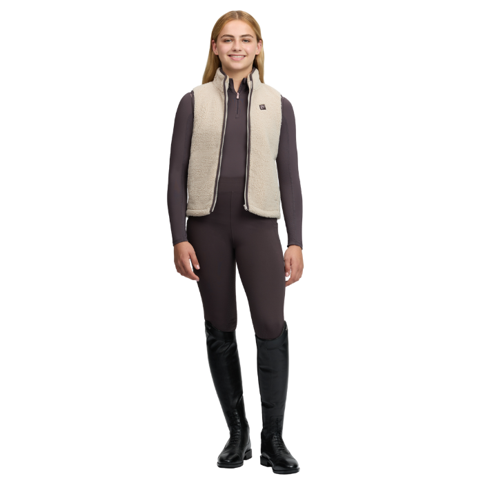 LeMieux AW26 Young Rider Trinity Teddy Gilet Varient Almond. A woman modeling a beige sherpa fleece vest over a dark brown riding outfit, including a long-sleeve top, breeches, and tall black riding boots, against a plain white background.