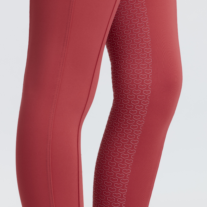 LeMieux AW26 Mini Pull Ons Varient Spiced Earth. Close-up of a person's leg wearing textured red athletic leggings with a white geometric pattern on the side, set against a light gray background.