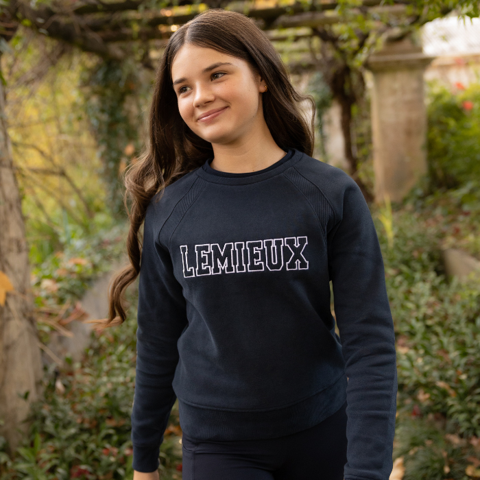 LeMieux AW26 Young Rider Shay Varsity Crew Varient Navy. A young woman with long brown hair stands outdoors wearing a dark navy crewneck sweatshirt with 'LEMIEUX' printed across the chest in white outlined block letters, paired with matching dark pants.